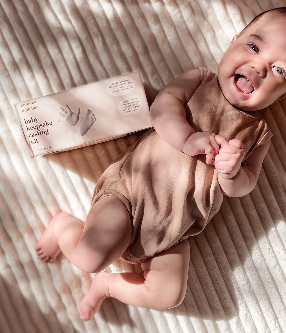 Baby in a pink onesie lying on a striped blanket next to a baby keepsake casting kit.